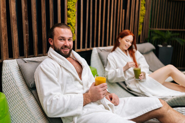 Portrait of cheerful bearded man and redhead woman in white bathrobes sitting on loungers by poolside, enjoying drinking fresh fruit cocktail at spa salon, after massage treatment, looking at camera.