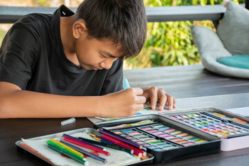 A boy in casual black clothes is practicing drawing and painting by using small watercolor rectangle bars in a paint box on a table on terrace of his summer vacation.