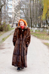 red-haired woman wearing white knitted hat and dressed in warm hooded casual parka jacket outerwear, girl in the parking lot, pedestrian crossing and car in the background, girl warms frozen © Oleksandra