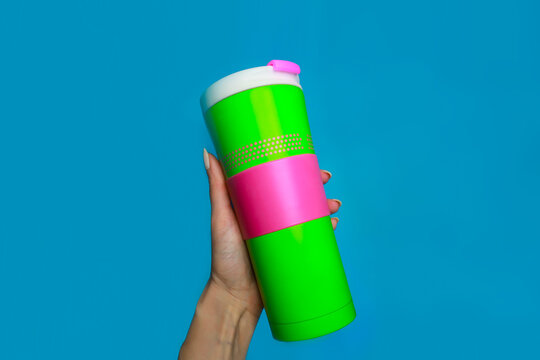 Woman's hand holding a bright reusable eco-friendly mug. Green cup with hot pink rim isolated on blue background. The lid is white. Suitable for tea and coffee.
