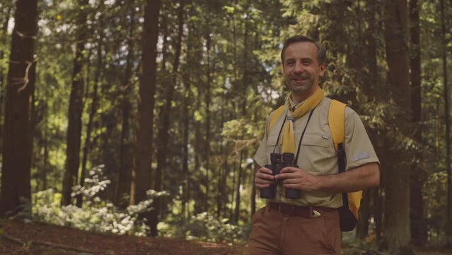 Medium portrait of mid aged Caucasian male scout leader with binoculars posing for camera in sunlit green summer forest