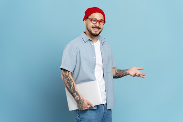 Authentic portrait of smiling confident asian man, freelancer holding laptop, isolated on blue background. Korean hipster wearing stylish eyeglasses, red hat looking at camera. Successful business