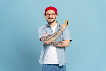Successful smiling Asian man wearing red hat, stylish eyeglasses, holding credit card looking at camera isolated on blue background. Happy confident freelancer receive payment. Shopping, sales concept