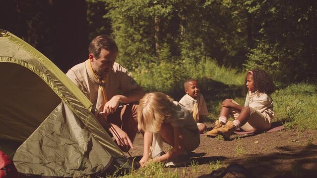 Little Caucasian boy scout and adult scouter setting up tent together on forest glade lit by sunlight