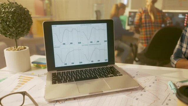 POV shot of the man putting down a paper cup of coffee and discussing a new project or report on a laptop screen with a young manager. The youth team is working on computers in the background.