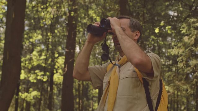 Medium shot of adult Caucasian male scouter in uniform looking through binoculars while spending time in summer forest