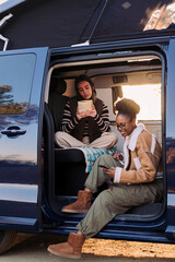 two young women enjoying camper van life reading at sunset, concept of travel adventure and female...