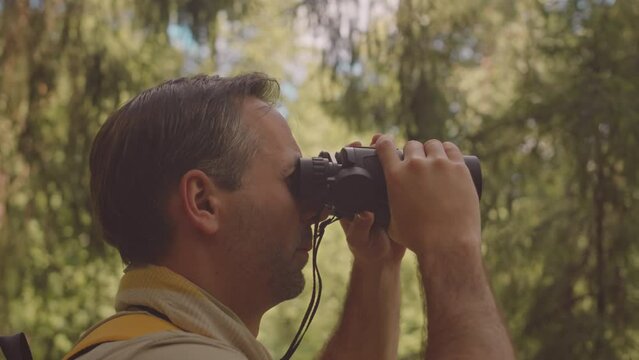 Adult Caucasian male scouter hiking and camping in summer forest, exploring area with binoculars