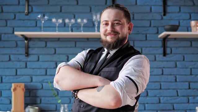 Behind the bar counter, a charming and charismatic bartender with a big smile folds hands and poses for the camera. The bartender makes eye contact with the camera. Flourishing restaurant enterprise.