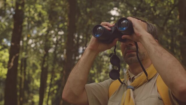 Medium shot of responsible adult Caucasian male scout leader in uniform discovering forest area through binoculars