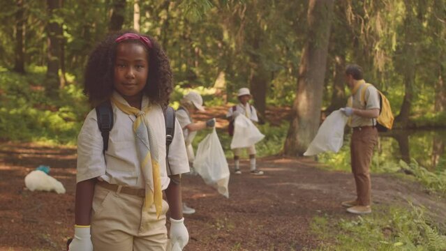 Medium portrait of little African American girl scout posing for camera in forest while her troop with scout leader in head collecting trash in bags at background