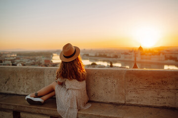 Naklejka premium Smiling female tourist in a hat admires the landscape of the city at dawn. Euro-trip. Travel, tourism and active lifestyle concept.