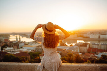 Smiling female tourist in a hat admires the landscape of the city at dawn. Euro-trip. Travel,...