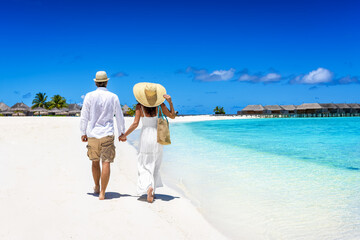 Happy couple in white summer clothing and with hats walks on a tropical paradise beach with turquoise sea in the Maldives islands