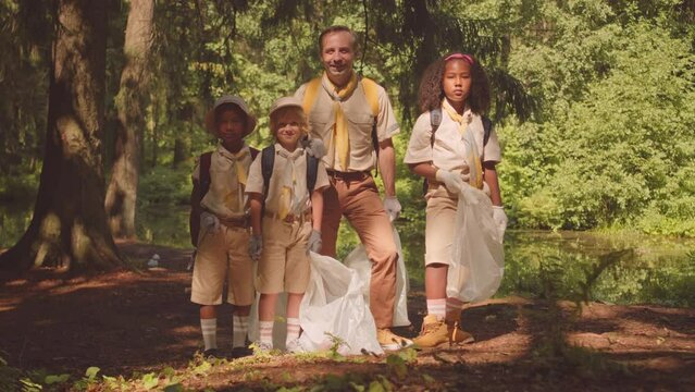 Full length slowmo portrait of adult Caucasian male scout leader with troop of multiethnic kids with rubbish bags posing for camera standing in sunlit green forest after collecting it from trash