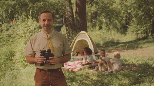 Medium slowmo portrait of smiling Caucasian male scouter with binoculars looking at camera standing on sunny forest glade with diverse cub scouts resting on grass in background
