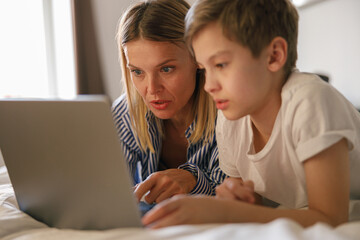 Small cute schoolboy with mother is using laptop in online learning at home while lying on bed