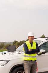 portrait engineer in reflective clothing and safety helmet next to a car in a field
