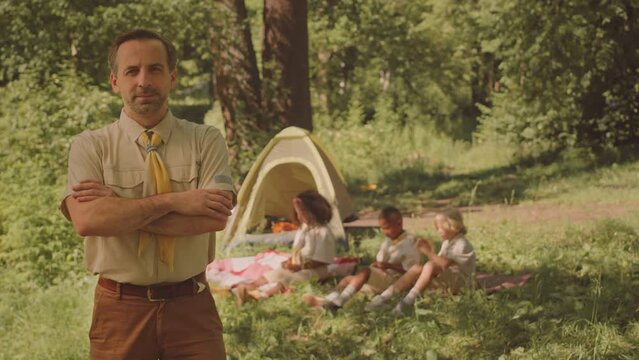 Medium slowmo portrait of confident mid aged Caucasian male scout leader posing for camera with hands folded while spending summertime with kids in forest camp
