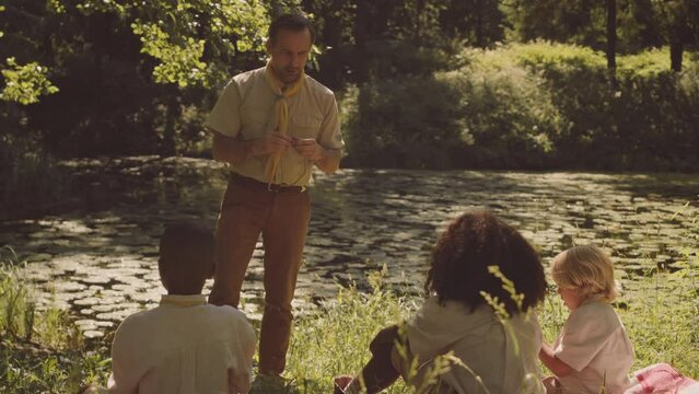 Mid adult Caucasian male scout teacher giving lesson of tie making to multiethnic group of kids sitting on green forest glade under sunlight camping together