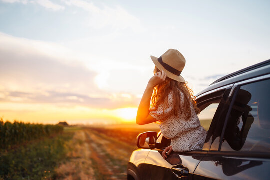 Young happy woman in hat enjoys car ride, leaning out of the window. In summer, a beautiful tourist woman travels by car and enjoys the golden sunset. Concept of an active lifestyle, travel, tourism.