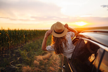 Naklejka na ściany i meble Young happy woman in hat enjoys car ride, leaning out of the window. In summer, a beautiful tourist woman travels by car and enjoys the golden sunset. Concept of an active lifestyle, travel, tourism.