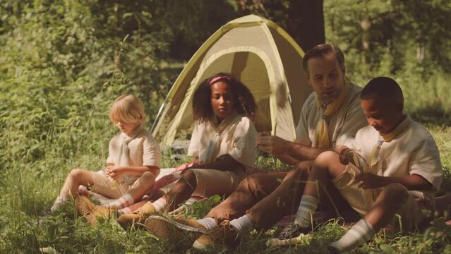 Adult Caucasian male scout leader teaching multiethnic group of kids to make ties while sitting on sunlit forest glade by camping tent