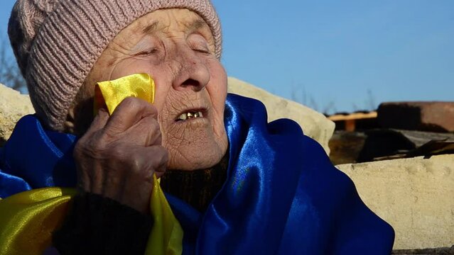 Grandmother in tears because of the war. War in Ukraine. Russian aggression against Ukraine.Peace concept. Elderly woman with Ukrainian flag. Destruction of Ukrainian cities. Ukrainian refugees. 