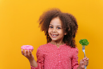 Studio shot of a smiling Black girl holding fresh broccoli and donut on a yellow background.