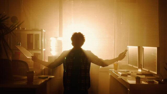 The silhouette of a man falls to his knees, raises his hands up and laughs in a dark office in front of a window with bright backlight. Enlightenment, insight, god of marketing.