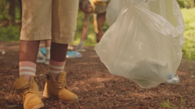 Low section of unrecognizable kid in scout uniform picking up plastic bottle from ground while cleaning up forest area from rubbish together with troop