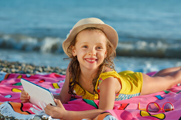 A happy child in a swiming suit and a summer hat lies on a bright blanket with a tablet in her hands.