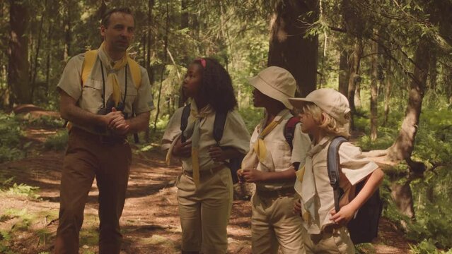 Medium shot of adult Caucasian male scout leader and group of multiethnic kids in scout uniform with backpacks exploring forest area together on sunny day