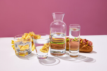 Still life with fast food hot dog sandwich, french fries and glasses with water on white table over pink studio background. Junk food
