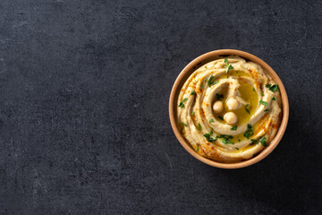 Chickpea hummus in a wooden bowl garnished with parsley, paprika and olive oil on black slate background. Top view. Copy space