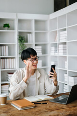 Attractive happy young Asian student studying at the college library, sitting at the desk, using a laptop, tablet and headphones having a video chat.