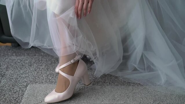 The bride in a white long luxurious wedding dress puts on high-heeled shoes.
