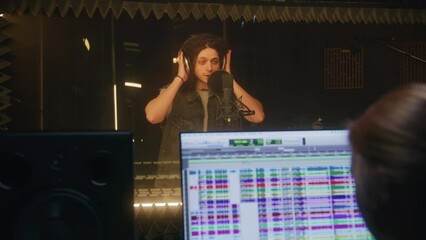 Vocalist comes in soundproof room and sings lyrical composition into microphone. Experienced sound engineer works in audio recording studio. Computer screen showing the DAW software. Music production.