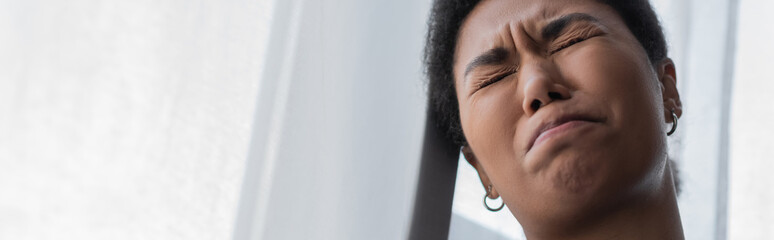 Low angle view of depressed multiracial woman standing near curtain at home, banner.