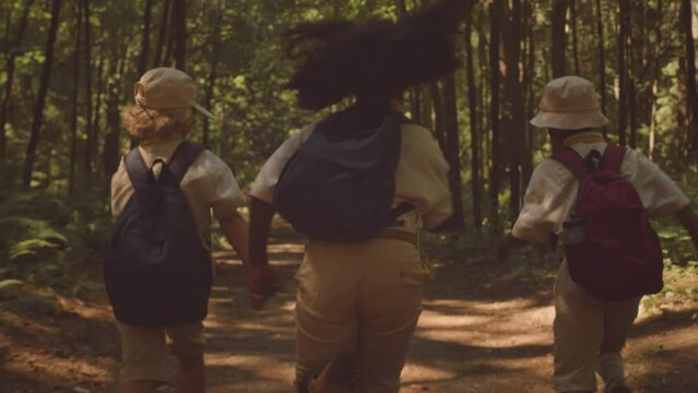 Slowmo back view of three multiethnic elementary age kids with backpacks running along sunny forest trail