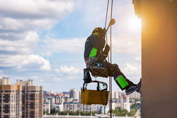 Professional industrial mountaineering worker washing exterior facade glazing hangs over residential facade building while at blue sky. Rope access laborer hangs on wall of skyscraper. Copy text space - obrazy, fototapety, plakaty