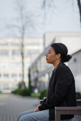 Side view of young multiracial woman with depression sitting on bench on urban street.