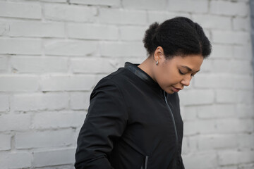 Depressed multiracial woman in jacket looking down on urban street.