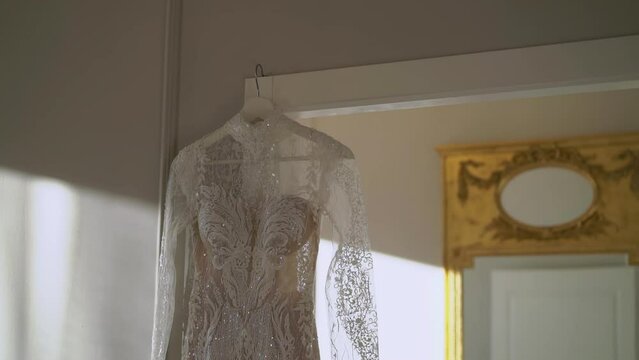 A beautiful white wedding dress hangs on a hanger at mirror in the bedroom. Morning of the bride, wedding day. Women's fashionable clothes for the holiday.