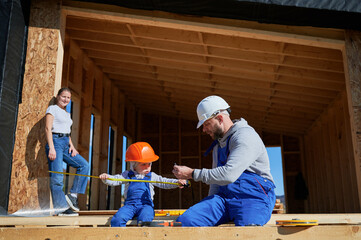 Father, mother and son building wooden frame house. Toddler boy helping his daddy, while woman...