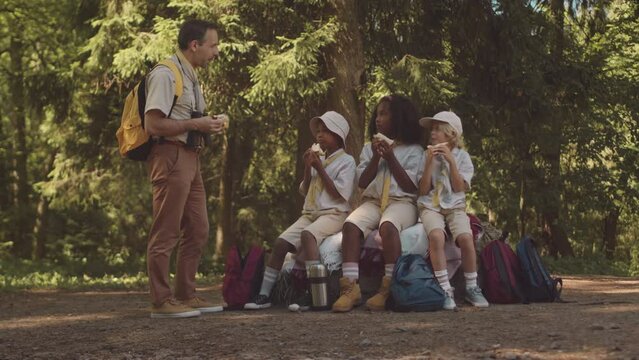Full length shot of diverse group of cub scouts sitting on their backpacks having lunch while hiking in forest together with their scout leader