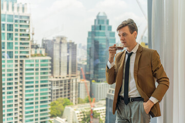 Fototapeta na wymiar Handsome young white businessman drinking a cup of coffee standing looking out the window view of city skyline in a business district area