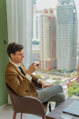 Handsome young white businessman holding a cup of coffee looking out the window view of city skyline in a business district area