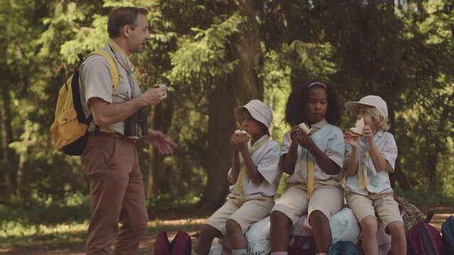 Medium shot of mid adult Caucasian male scouter with group of multiethnic elementary age kids having lunch while hiking together in forest