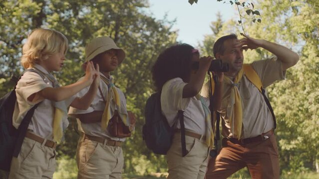 Medium shot of mid adult Caucasian male scouter teaching multiethnic group of kids to explore forest territory with help of binoculars